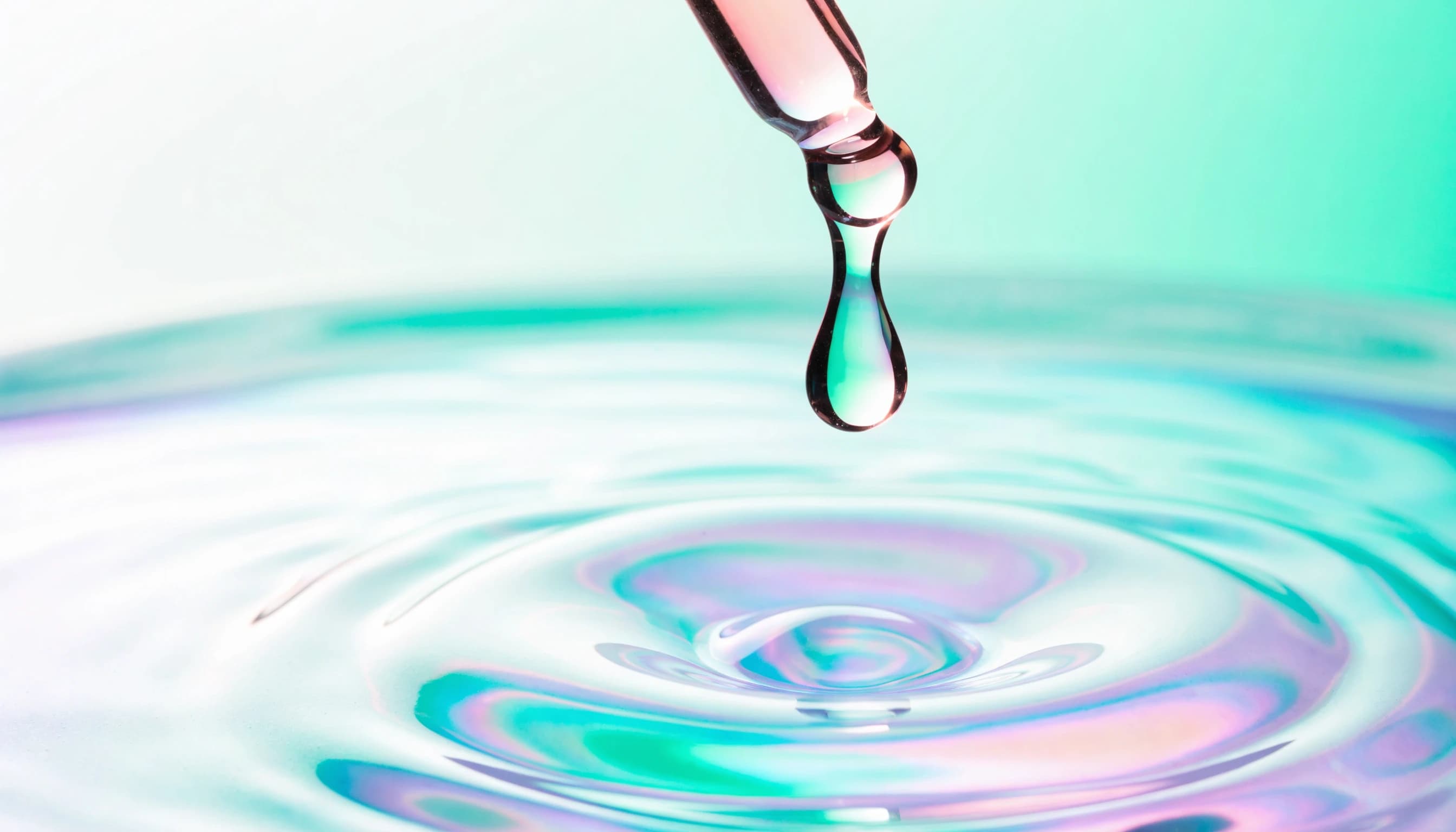 A drop of serum falling from a glass dropper into rippling iridescent liquid.